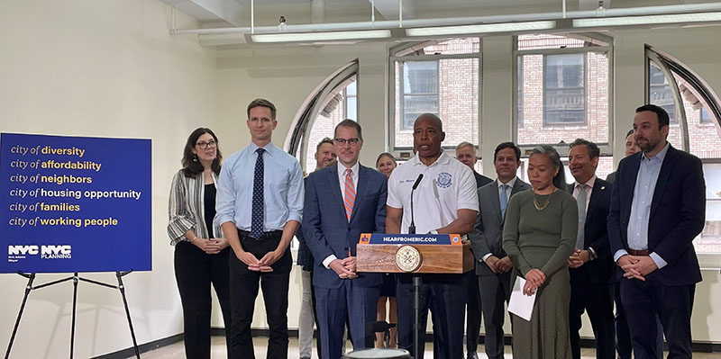 Photo of Mayor Eric Adams at a podium, with other NYC officials standing behind him and a sign that reads: “City of Diversity, City of Affordability, City of Neighbors, City of Housing Opportunity, City of Families, City of Working People  