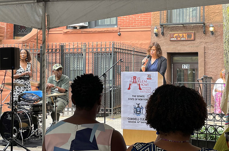 LPC Executive Director Lisa Kersavage speaks at a podium in front of a seated audience, a small band is visible in the left side of the photo