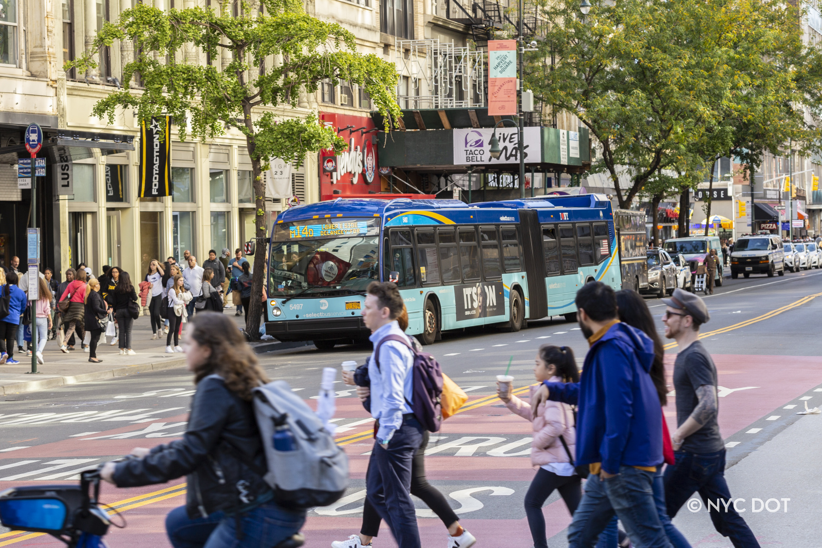 A blue MTA bus picks up passengers on 14th Street. Pedestrians cross the street. The roadway is painted red and marked with the text “Bus and Trucks Only”.