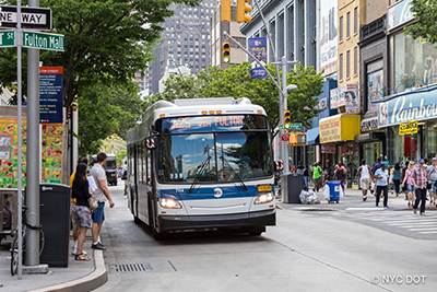 An MTA bus travels along Fulton Street in Brooklyn, while pedestrians walk on the sidewalk and wait at the curb to cross the street.