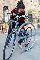A woman leans over to lock her bike to a round bicycle rack on a sidewalk.