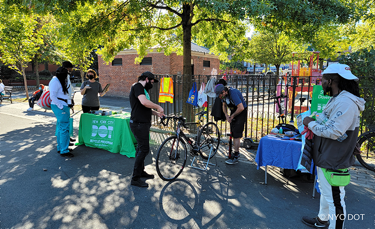 On a sunny day at Nostrand Playground in Brooklyn an NYC DOT employee speaks to two members of the public at a Biketober outreach event. Next to them, two men are repairing a bicycle, one of them is working on the breaks while the other is pumping air into the back tire. A fifth man stands watching the men repairing the bicycle.