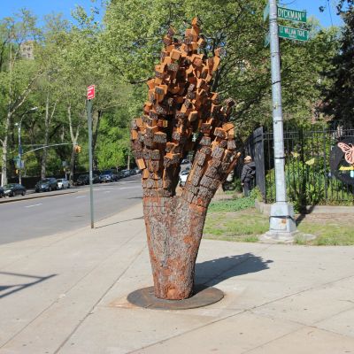 A sculpture shaped like a tree trunk with thick branches shaped into cubes installed on a wide sidewalk in N Y C.