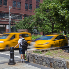 A pedestrian waits on a refuge island on a sunny day, while yellow taxis speed down the street which has a speed camera angled at the roadway.