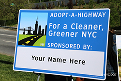Blue Adopt a Highway sign being presented on an easel. Sign reads: For a Cleaner Greener NYC Below that is a blank white space with the message  Your Name Here