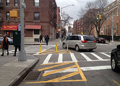 A car turns left around a slow turn wedge treatment, helping the driver to see a pedestrian walking in the crosswalk. Location is 89 Ave & 164 St in Queens, NY.