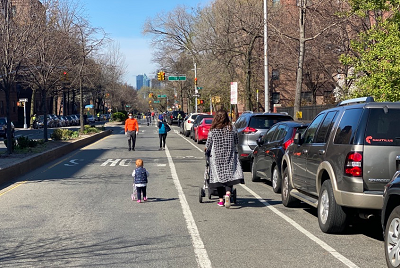 A mother and young child push strollers while walking on a street open to pedestrians and cyclists, while closed to vehicular traffic. Other pedestrians wearing face masks are walking in the distance