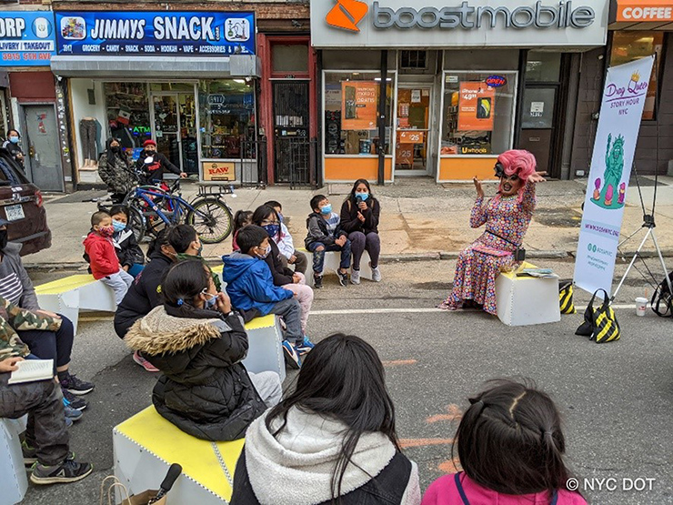 A colorful drag queen reads a story to children seated in a semi-circle on an Open Street in Sunset Park, Brooklyn.