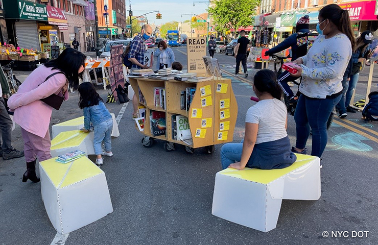 A pop up library on an Open Street is surrounded by yellow and white seating elements. Several passerby of varying ages stop to check out the books.