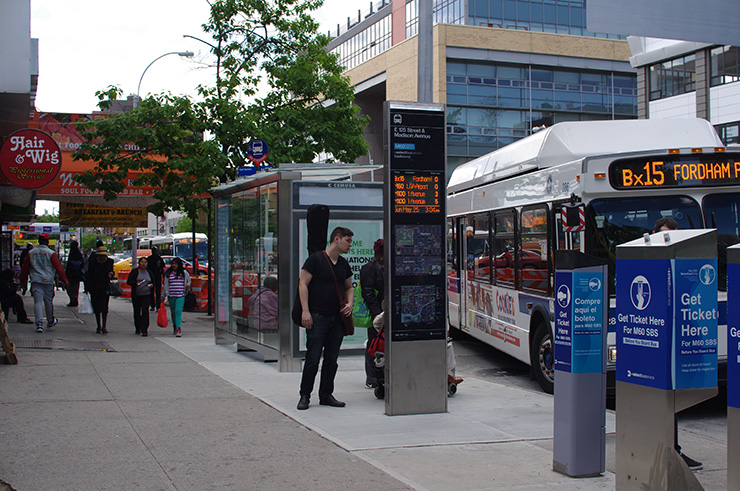At a bus stop, people look at a tall sign with maps and a digital display of arrival times for buses