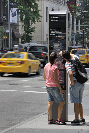 Two people look at a map on a tall, narrow sign on a sidewalk, while yellow taxis drive by