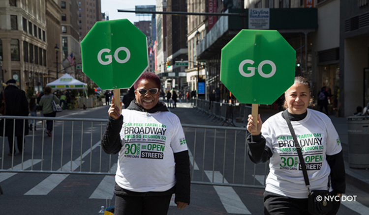 Two people in Car Free Earth Day shirts are standing on Broadway in Midtown Manhattan. They are holding green signs that day “GO” to direct pedestrians and cyclists.