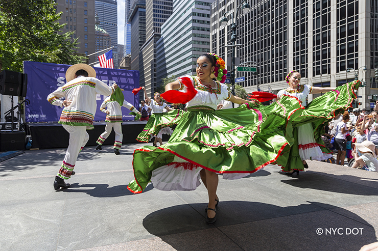 Woman and men dressed in bright green and white outfits dance on an outdoor stage in Manhattan