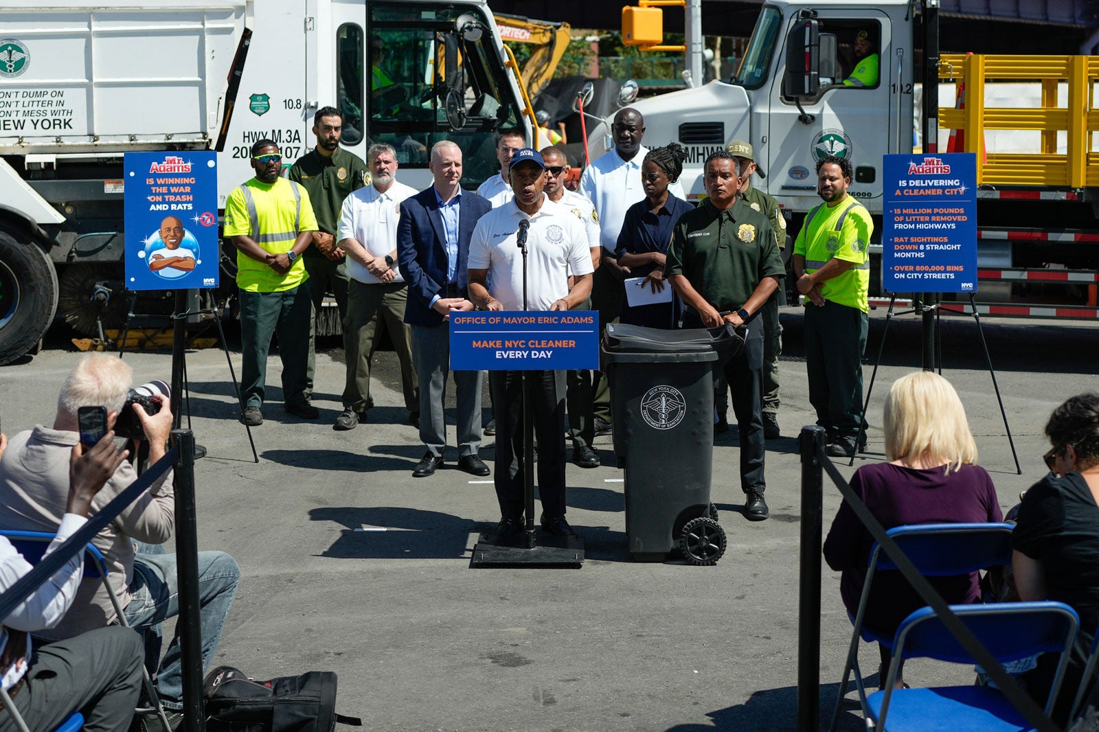 Mayor Adams speaks at the podium while a group of city employees and trash collect trucks were behind him.
