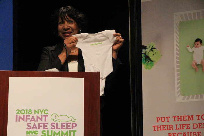 A woman holds up a baby onesie while standing on a podium with the 2018 N Y C Safe Sleep Summit logo on it