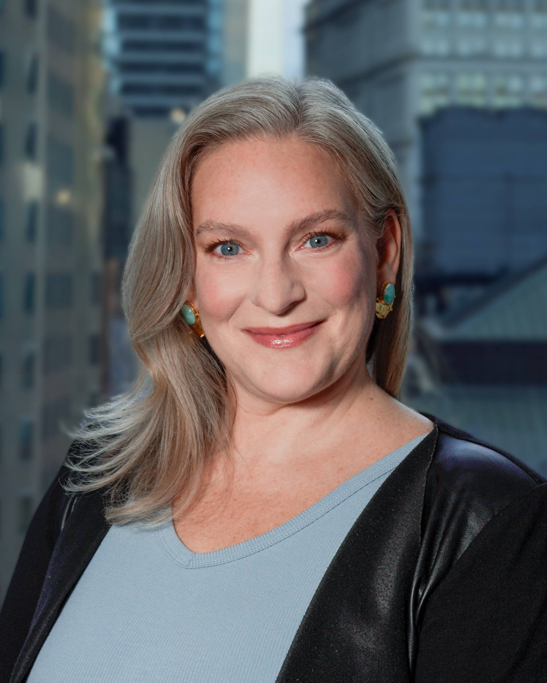 Portrait of Elizabeth Wolkomir in a black jacket, lightly smiling for a photo with a backdrop of city buildings