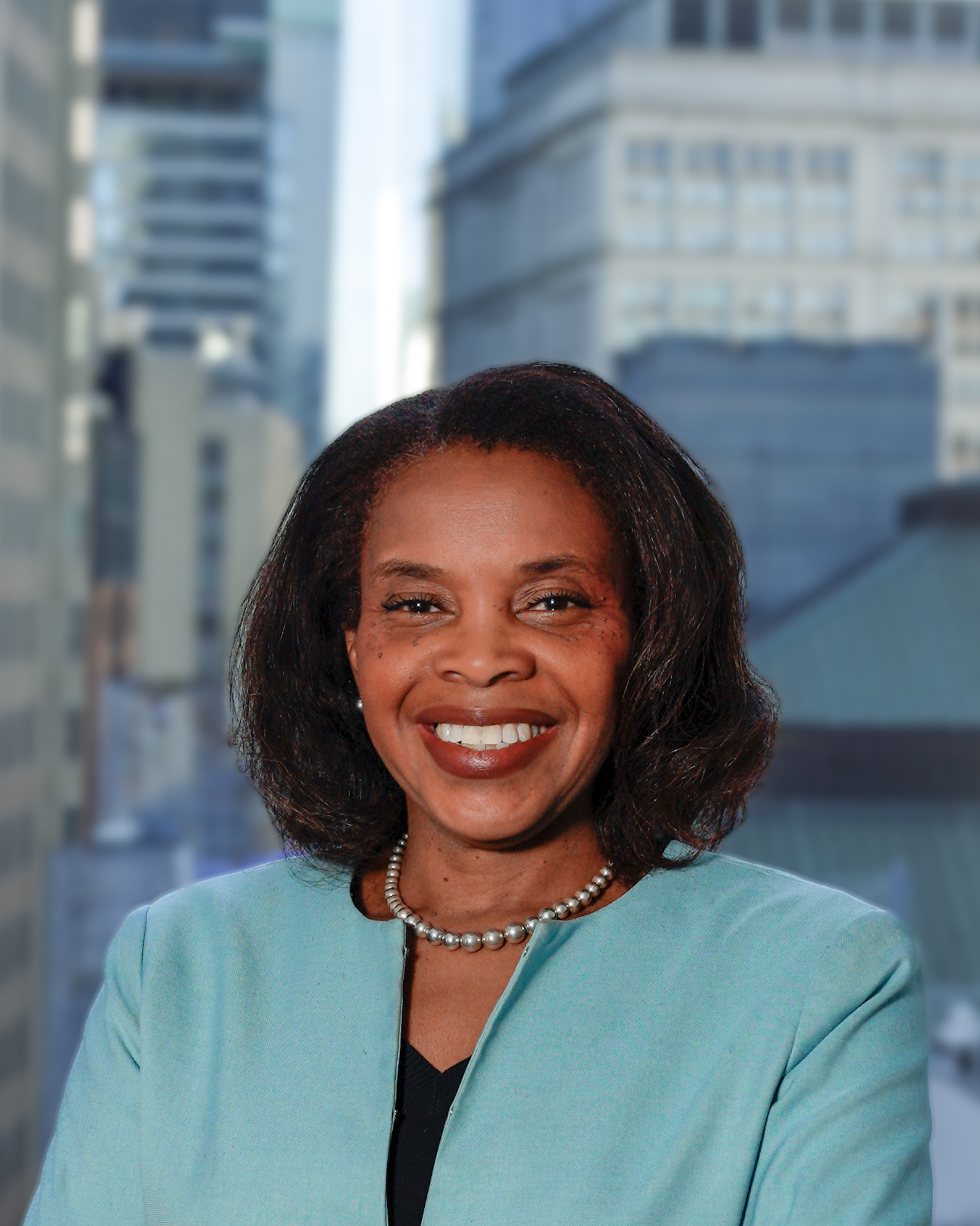 Portrait of Ina Mendez in a blue jacket and a beaded necklace, smiling for a photo with a backdrop of city buildings