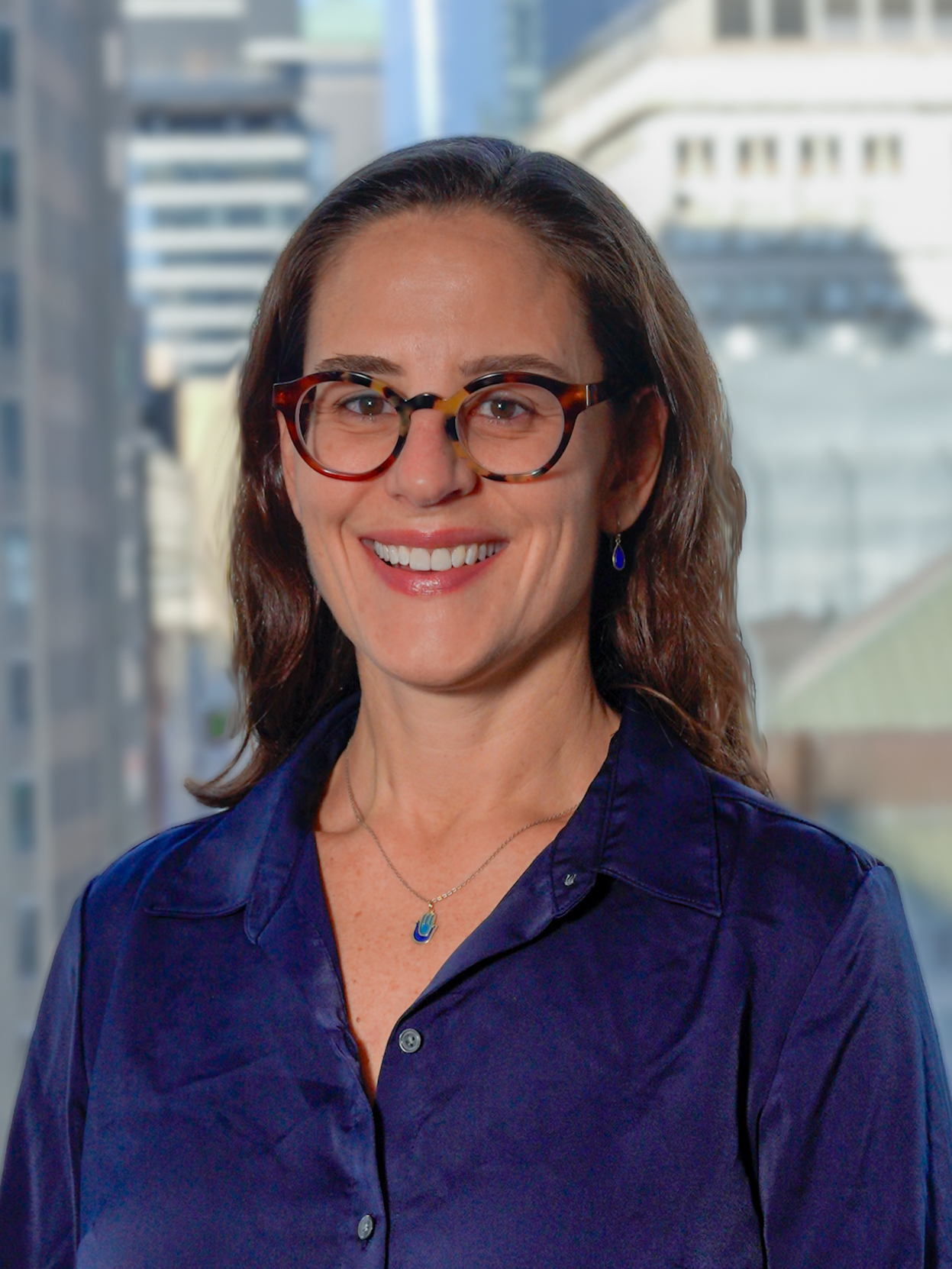 Portrait of Jill Krauss wearing glasses and a blue shirt as she is smiling for a photo with a backdrop of city buildings