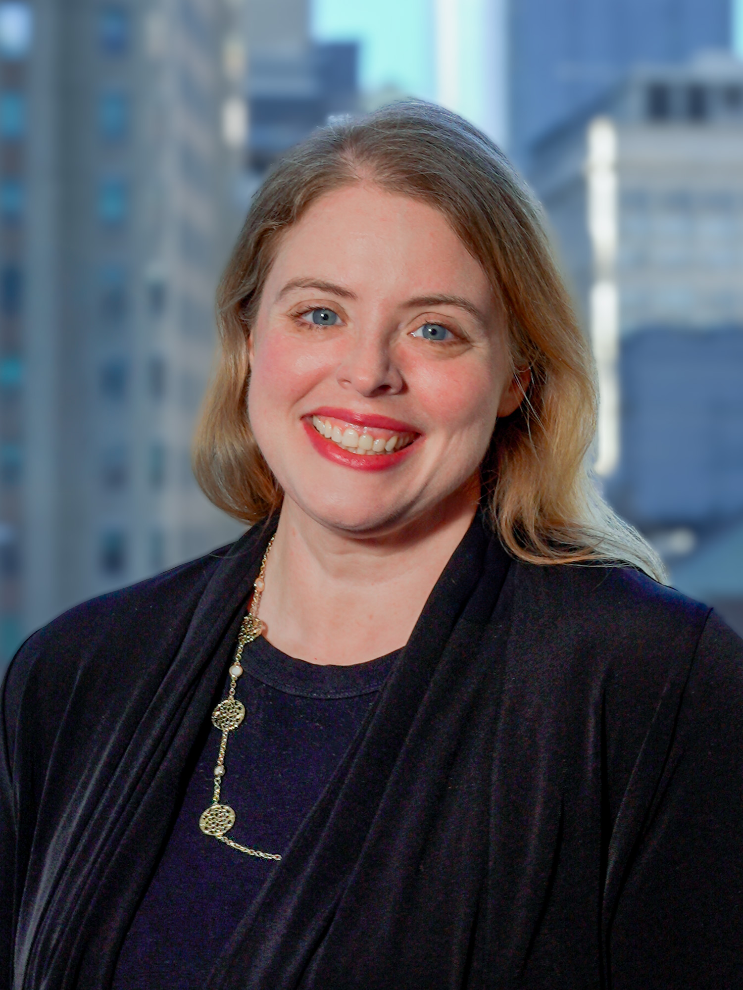 Portrait of Joanna Rose in a black jacket and a necklace, smiling for a photo with a backdrop of city buildings