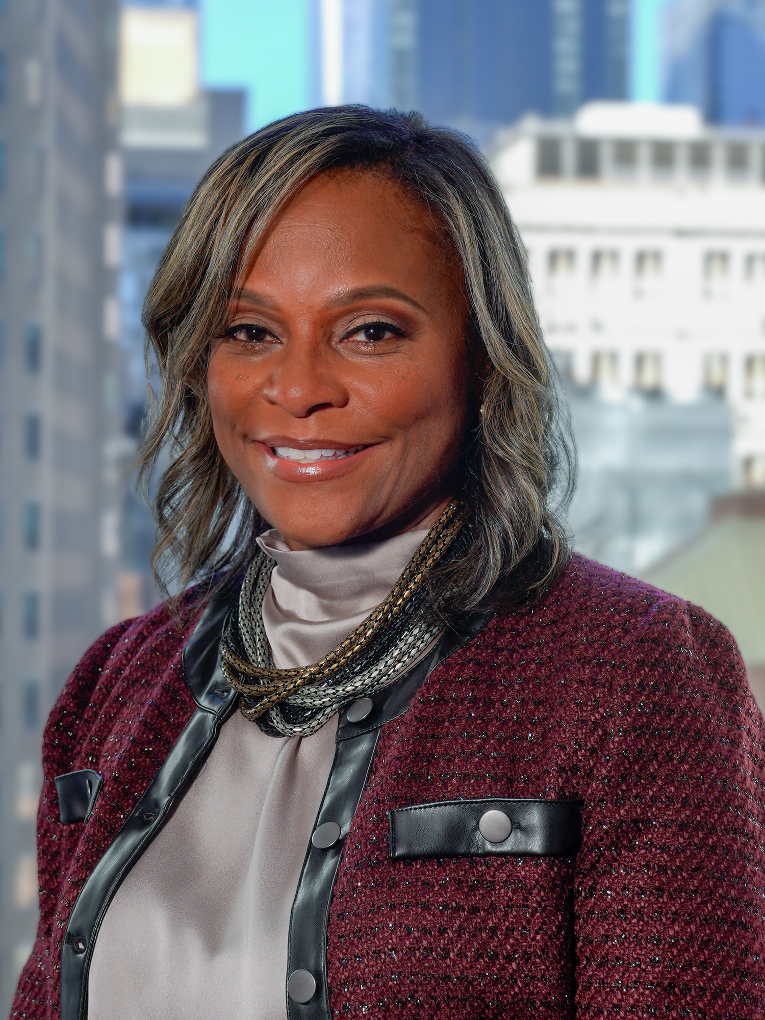 Portrait of Lisa Hines-Johnson in a purple jacket/blazer wearing many necklaces and smiling for a photo with a backdrop of city buildings