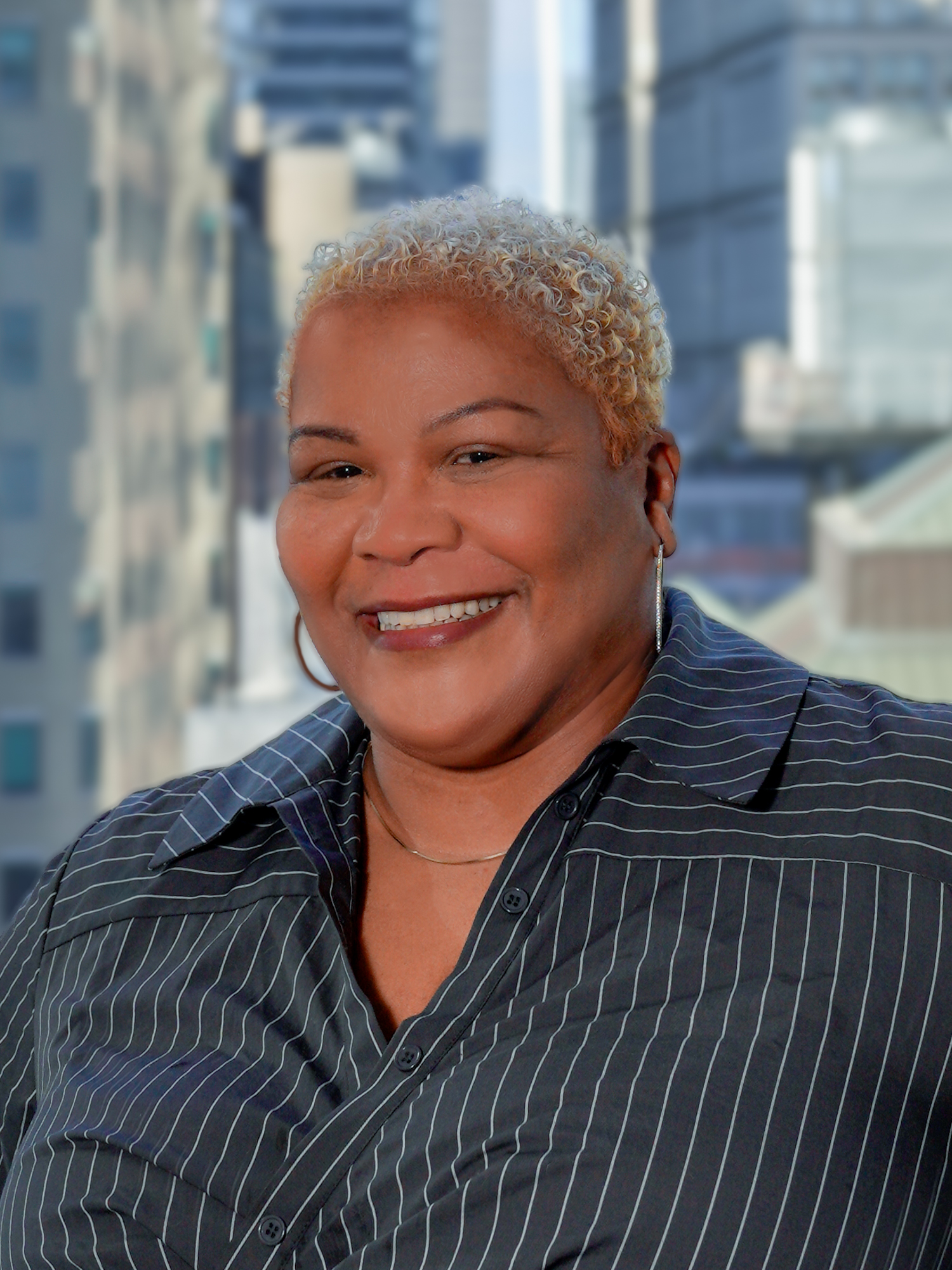 Portrait of Luisa Linares in a black collared shirt, smiling for a photo with a backdrop of city buildings