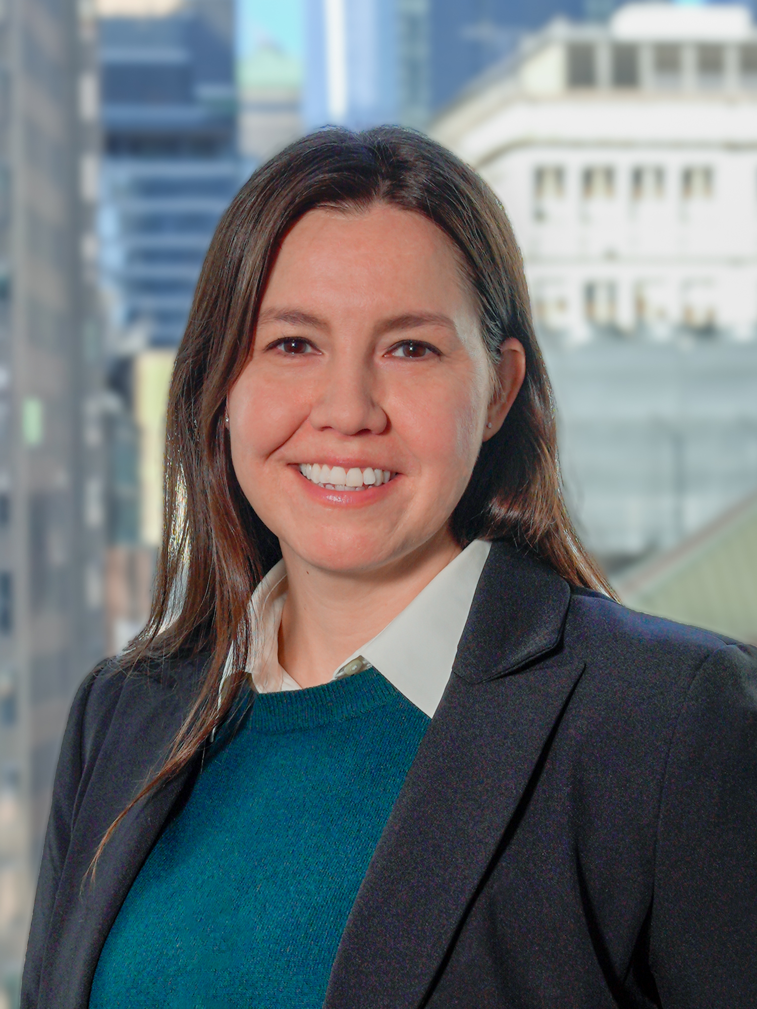 Portrait of Margaret Pletnikoff in a black blazer and a blue sweater smiling for a photo with a backdrop of city buildings