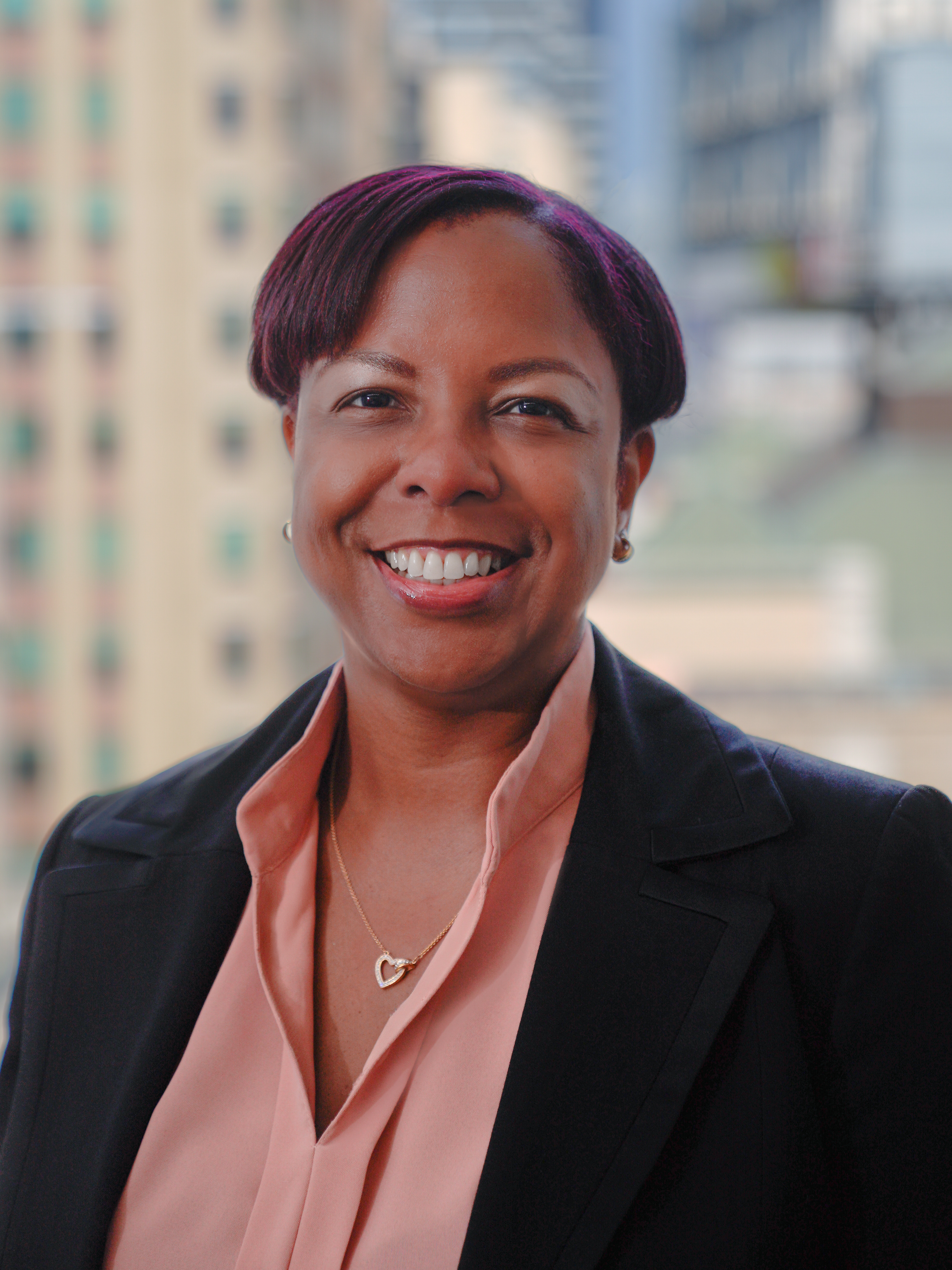 Portrait of Nikisha Ferguson wearing a black blazer and a pink shirt, smiling for a photo with a backdrop of city buildings