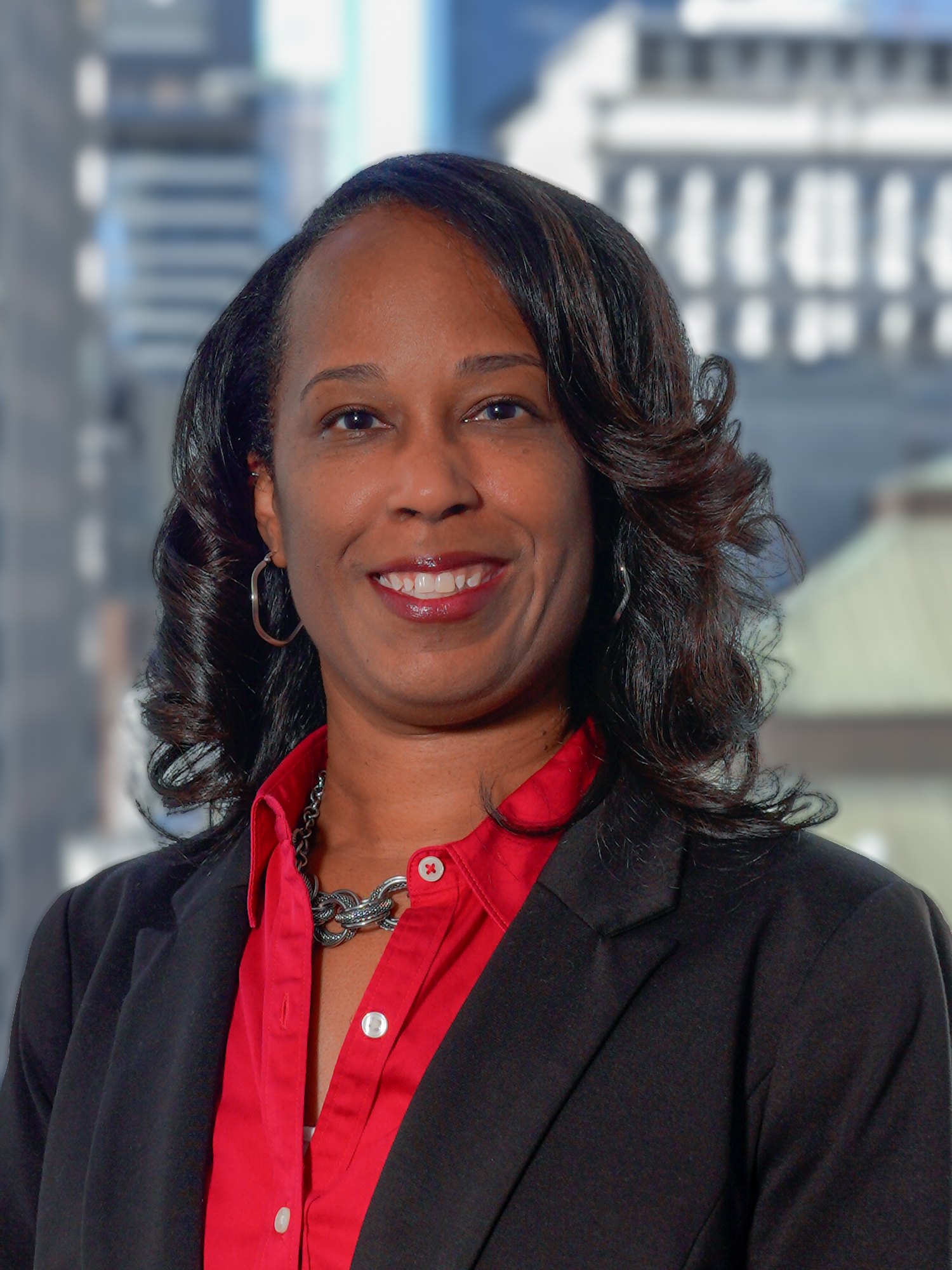 Portrait of Tia Waddy in a blazer and a button down shirt, smiling for a photo with a backdrop of city buildings