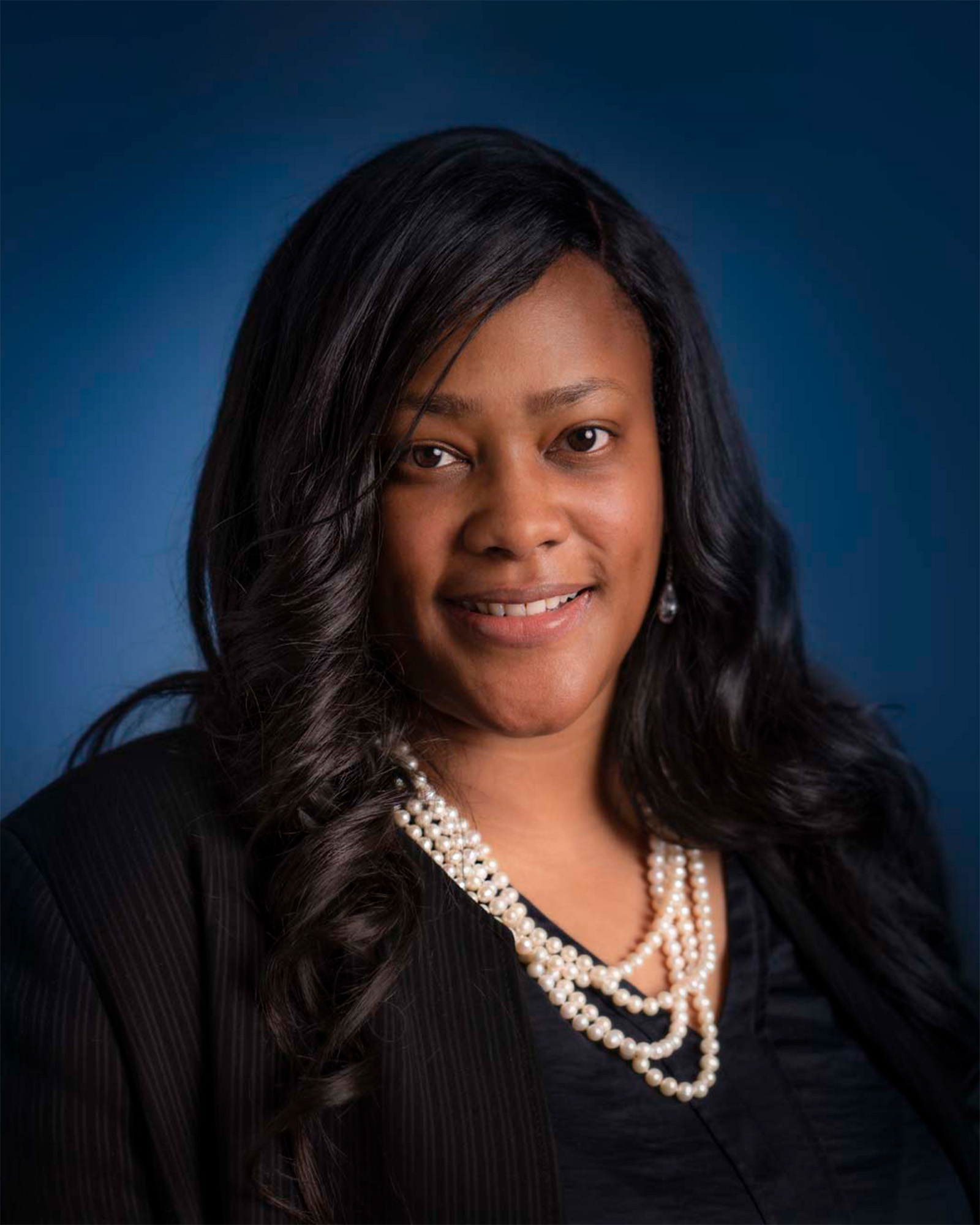 Portrait of a Winette Saunders in a black suit, smiling for a photo and wearing a beaded necklace