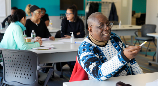 Learners in a Workforce Institute classroom: a gentleman asks a question, while a group at a table behind him discusses the training.
