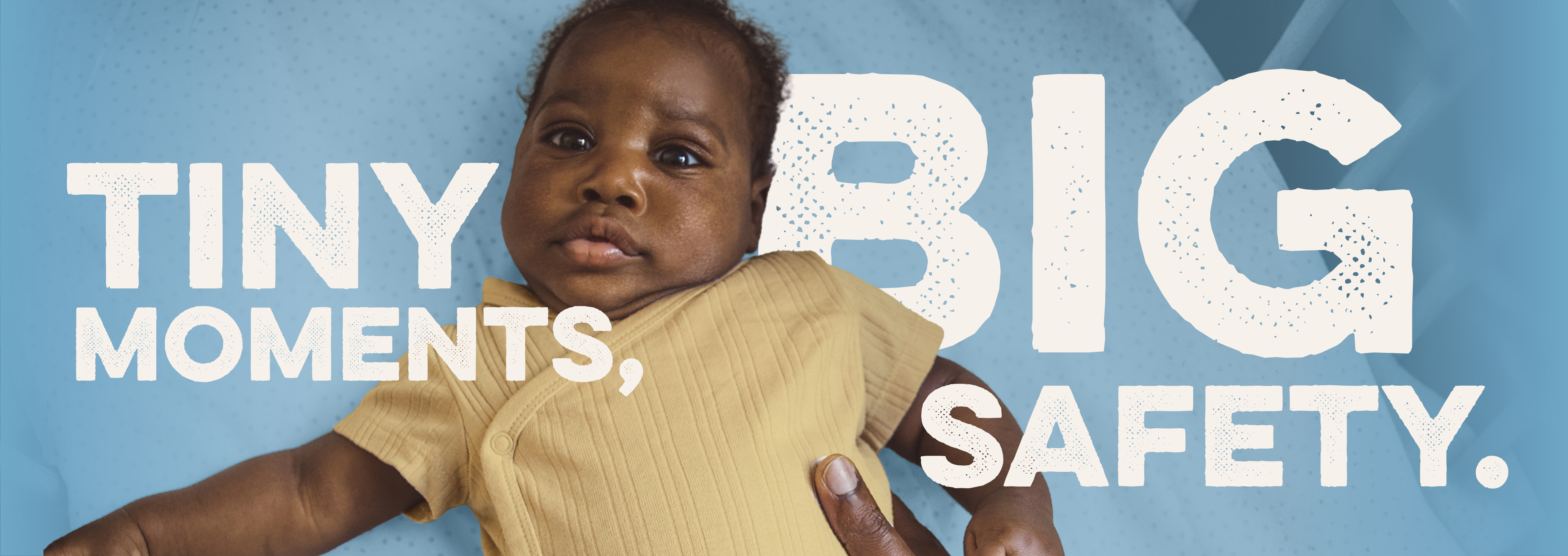 A dark-skinned baby is laying down and looking at something that is off camera. The baby is wearing a yellow onesie. An adult-sized hand is on the baby's stomach area. White text on a blue background reads 'Tiny Moments, Big Safety'