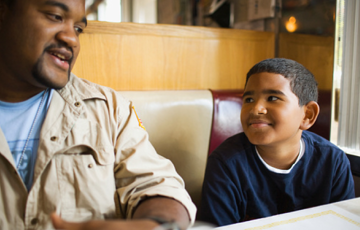 Parent and child sitting at a table as the son listens to his parent speak
                                           