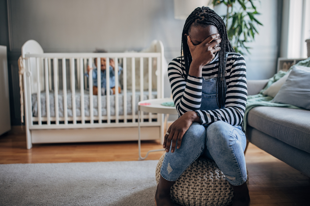 woman sitting with a hand to her face crying, background is a baby in a crib
                                           