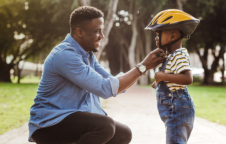 A father is putting a bicycle helmet on his young child. They are outdoors.
                                           