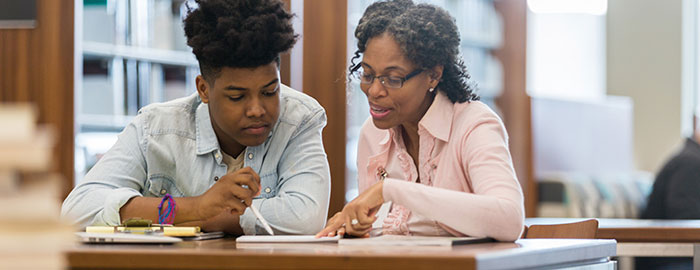 Two people sitting at a table reading a book and writing to it. One person is wearing glasses with a pink shirt, and the other is wearing a jean shirt and has a pen to the book