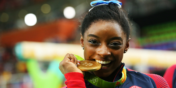 Photo of Simone Biles smiling and biting her gold medal
