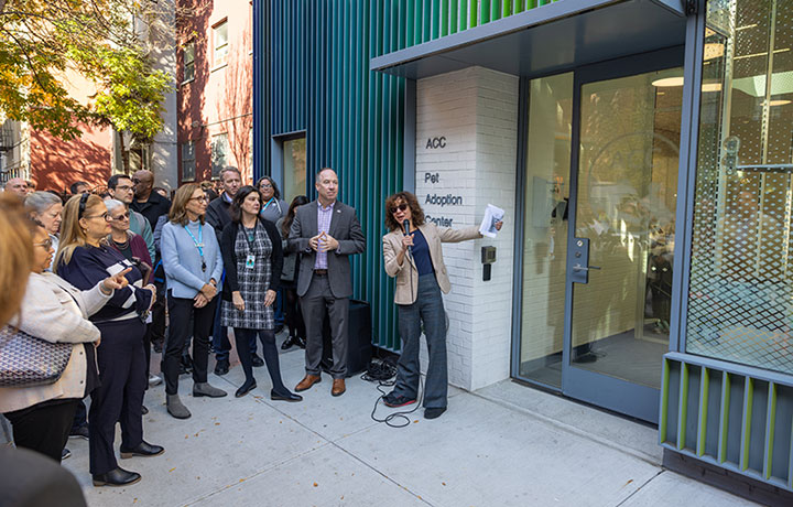 Photo of the ribbon-cutting of the Paul A. Vallone Queens Animal Care Center. Paul Vallone’s family members, city officials, and ACC representatives stand by the ribbon outside the entrance as others look on.
                                           