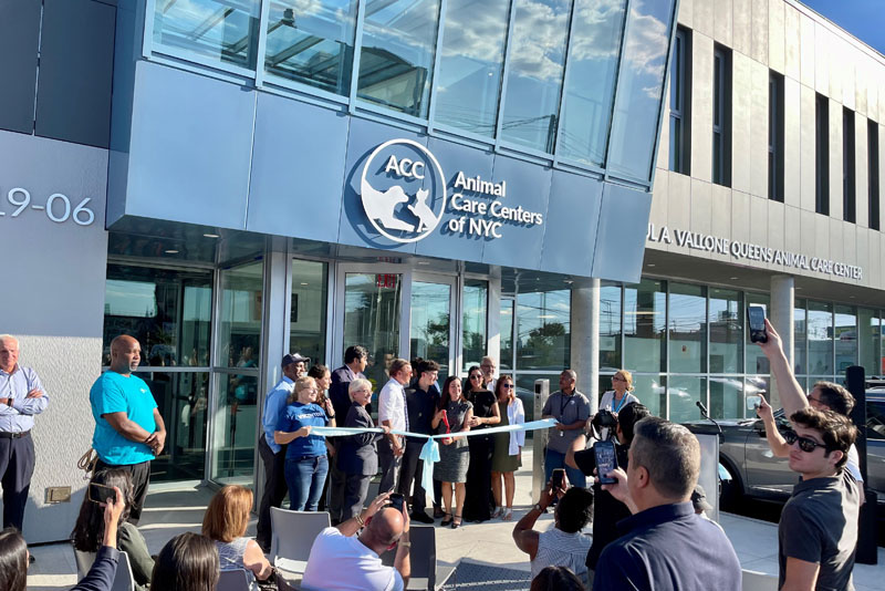 Photo of the ribbon-cutting of the Paul A. Vallone Queens Animal Care Center. Paul Vallone's family members, city officials, and ACC representatives stand by the ribbon outside the entrance as others look on.