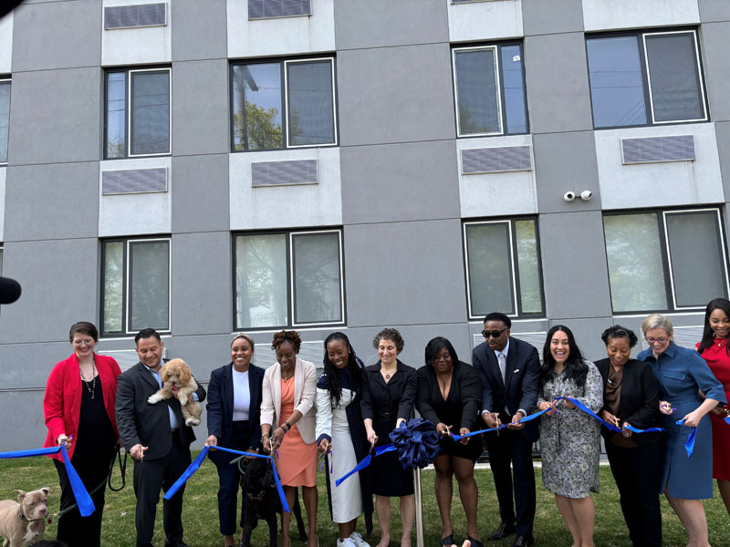 Photo of leaders of the Urban Resource Institute, DSS and DHS, and the community—some with dogs—cutting the ribbon on a new transitional housing facility.
