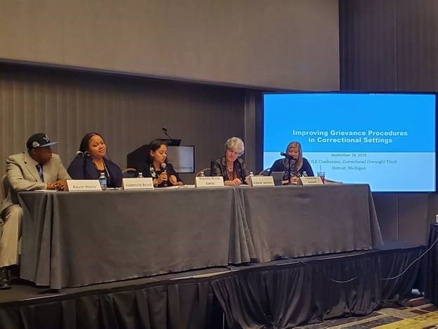 Five people on a sitting on a stage with a presentation screen that reads Improving Grievance Procedures in Correctional Settings