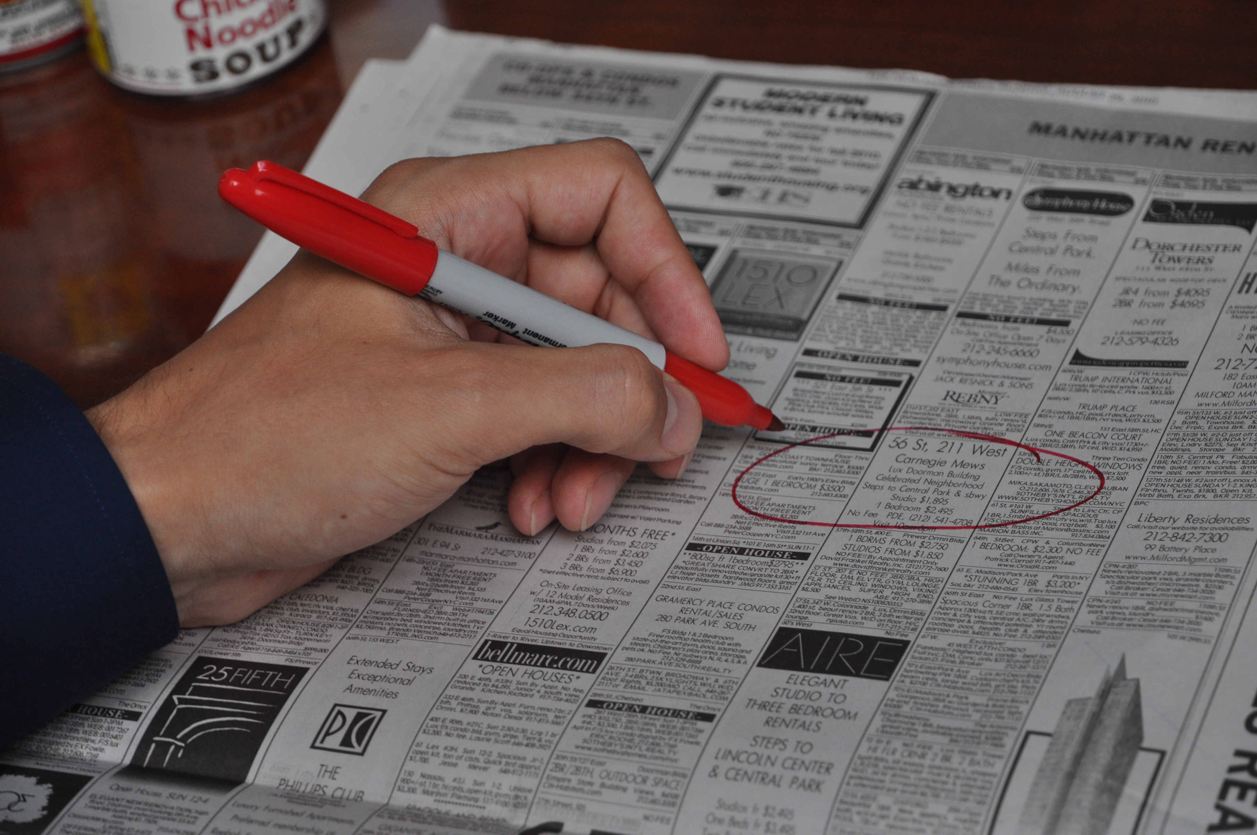 Hand holding  a red marker poised over the Housing Wanted section of a newspaper after  circling a specific ad