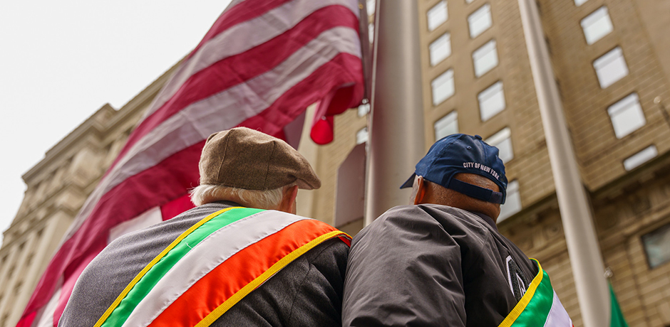 Mayor Adams stands alongside an honoree, both wearing decorative sashes, positioned in front of a flag pole during the Irish flag raising ceremony at Bowling Green park.