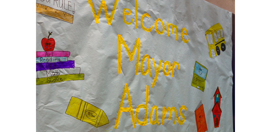 A white welcome sign with 'Welcome Mayor Adams' painted in yellow, decorated with colorful illustrations of educational items including books, crayons, a school bus, and a laptop.