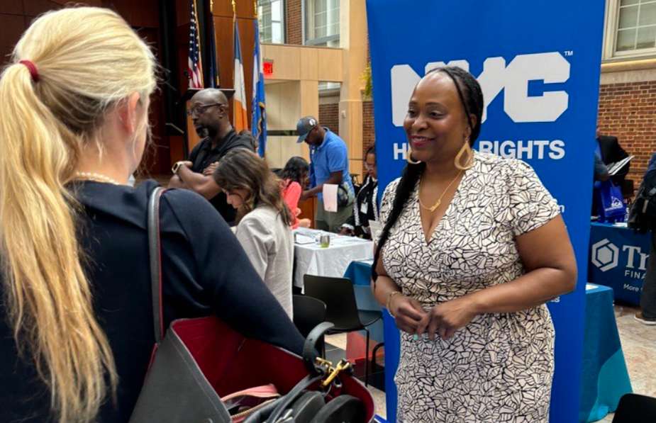 Two women speaking at an event with people and tables in the background. A banner with NYCCHR logo is also in the background.