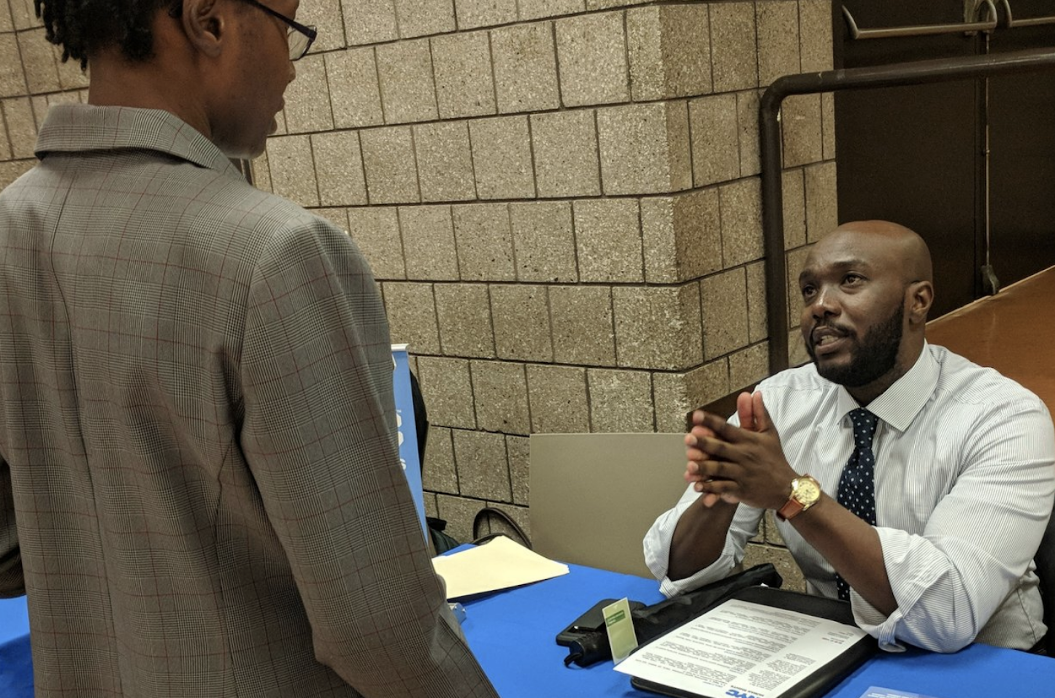 Woman speaking to a man who is seated behind a table.