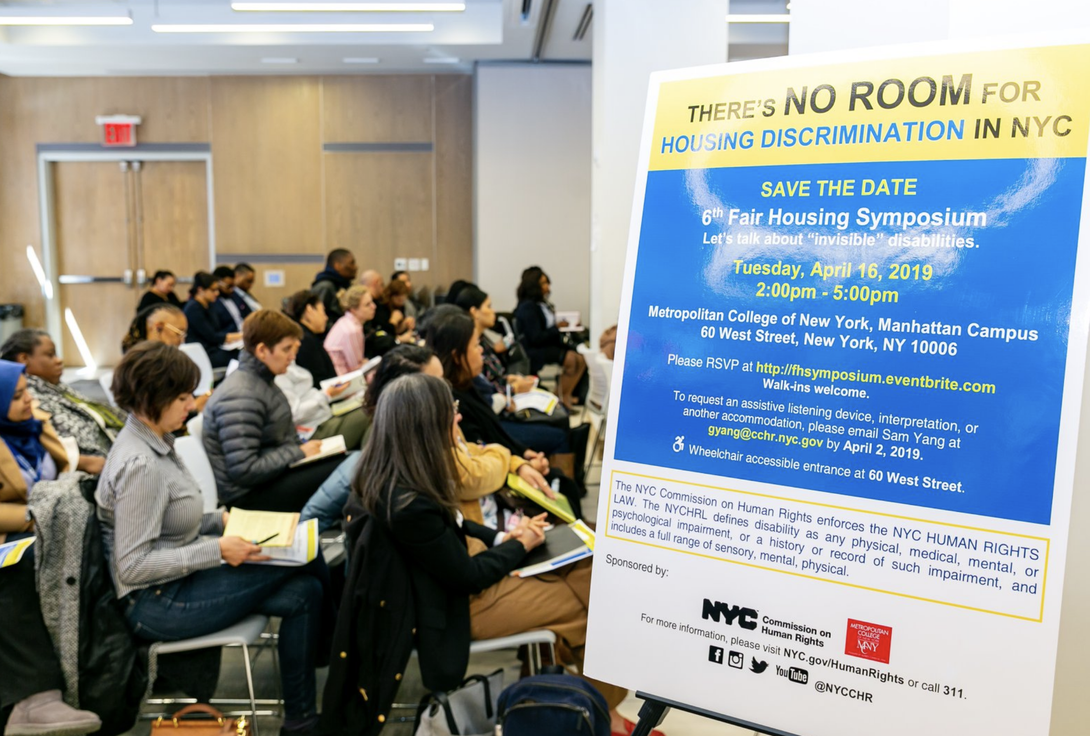 Profile of audience sitting at event next to a sign that reads: “There’s No Room For Housing Discrimination in NYC,” 6th Fair Housing Symposium, Tuesday, April 16, 2019.