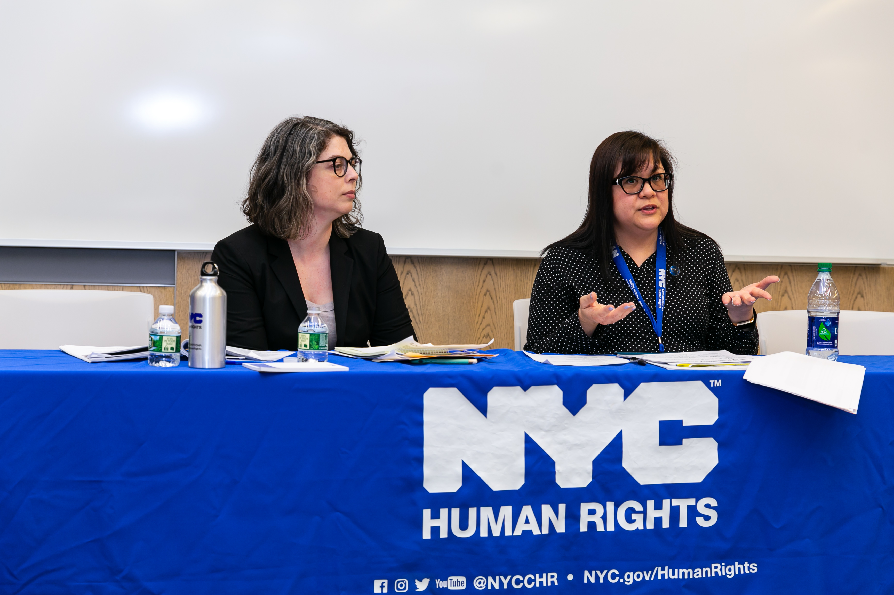 Two members of Commission staff sitting at table; one staffer is gesturing and speaking. Tablecloth reads, “NYC Human Rights.”