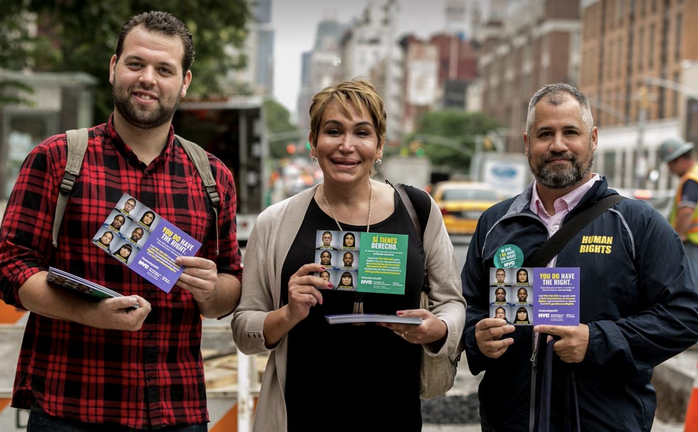 Three people, facing camera, holding up Commission palm cards that read “You Do Have The Right” in English and “Sí Tienes Derecho” in Spanish.