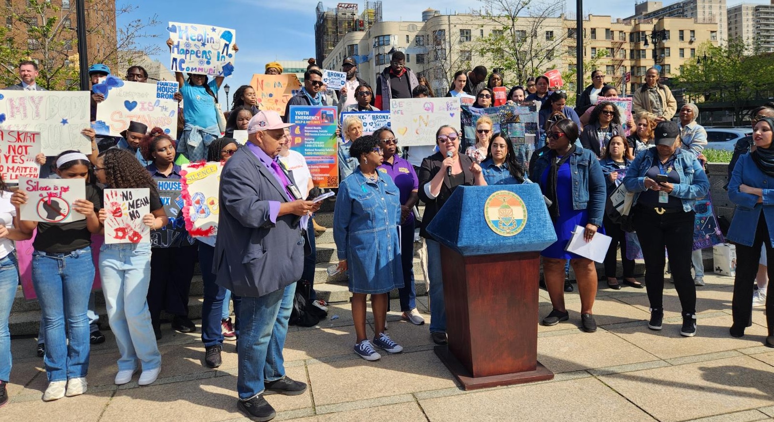 A group of people gathered outdoors at a public rally or community event, standing around a podium with a city seal on the front. Commissioner Christine Clarke is speaking into a microphone while others hold signs advocating against violence and supporting community safety and youth empowerment. Visible signs include messages such as “Healing Happens in Community,” “Silence is not YES,” and “No Means No.” The crowd includes adults and young people, with buildings and trees in the background on a sunny day.