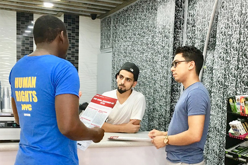 Man in NYC Human Rights shirt holds a brochure while talking to two business owners, one of which is standing behind a counter